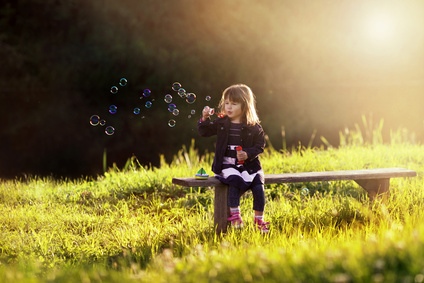 little girl sitting on a wooden bench blows bubbles in the rays