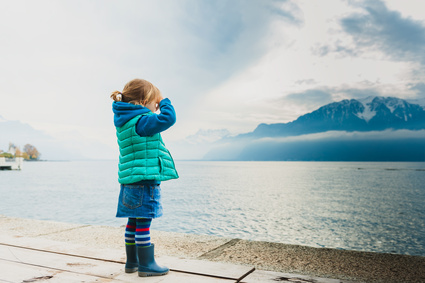 Adorable toddler girl playing by the lake, looking at the mountains, wearing, rain boots, jeans skirt and green waistcoat